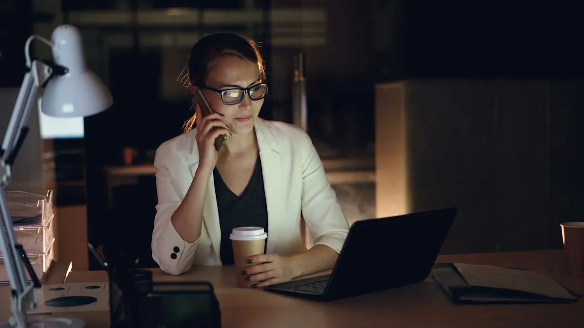 A coordinator on the phone at her desk late at night, with a laptop and a cup of coffee
